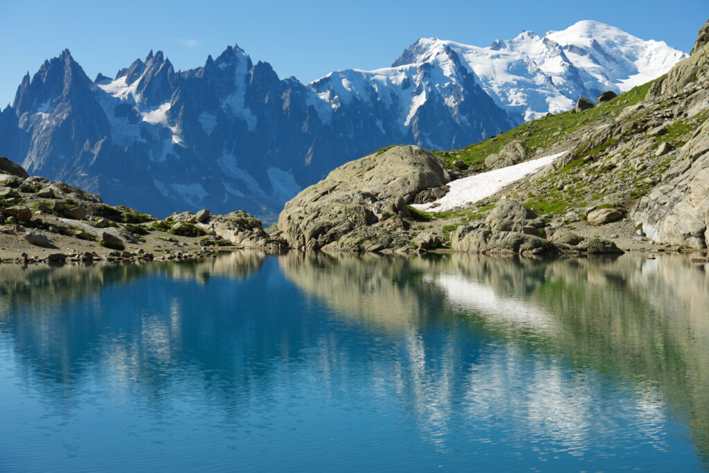 Lac alpin entouré de végétation verdoyante avec montagnes enneigées en arrière-plan sous un ciel bleu, typique du paysage savoyard.